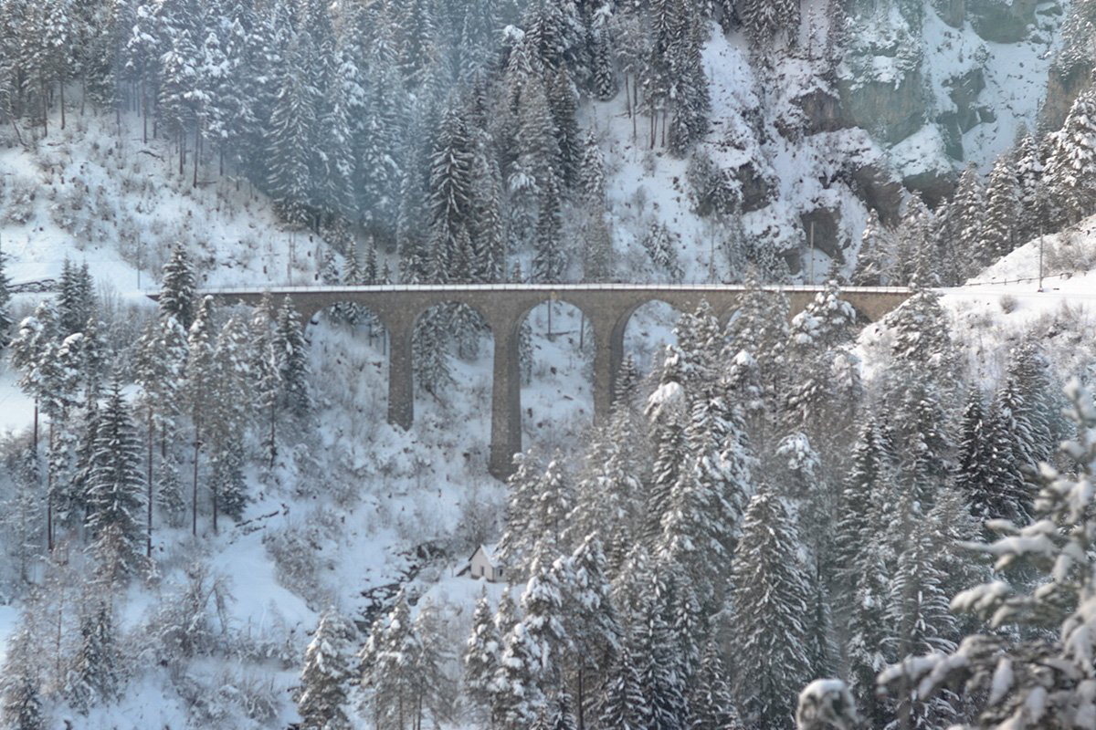 A snowy scene with a viaduct crossing two mountains.