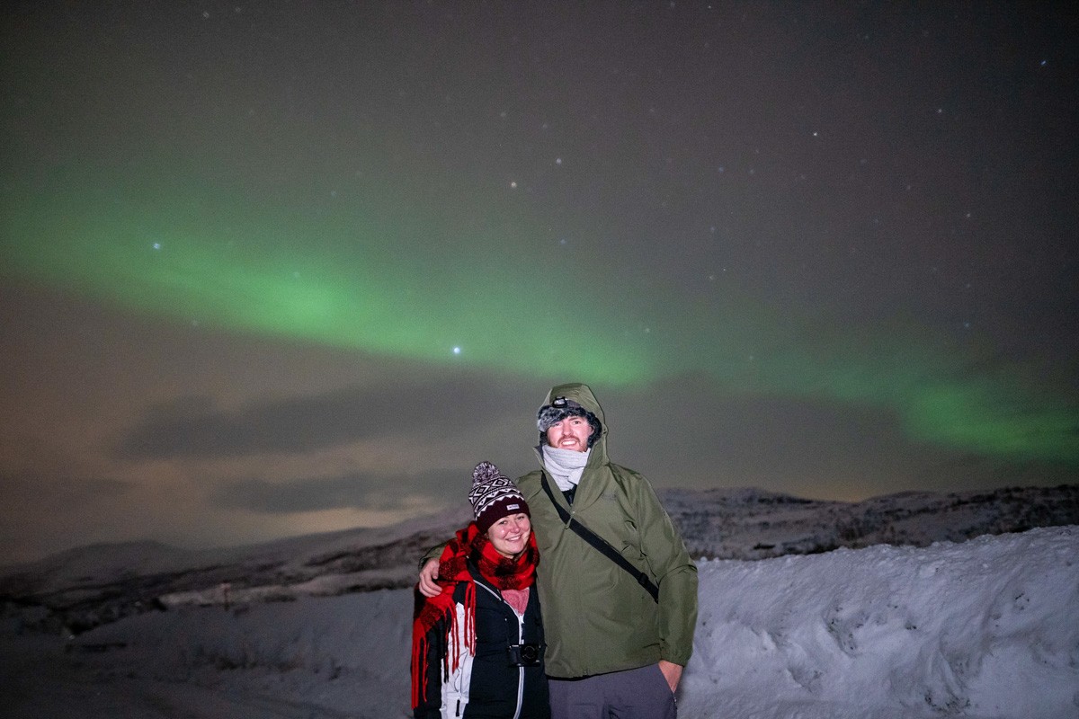 Claire and Richard standing in front of a snowy scene with the green hues of northern lights above