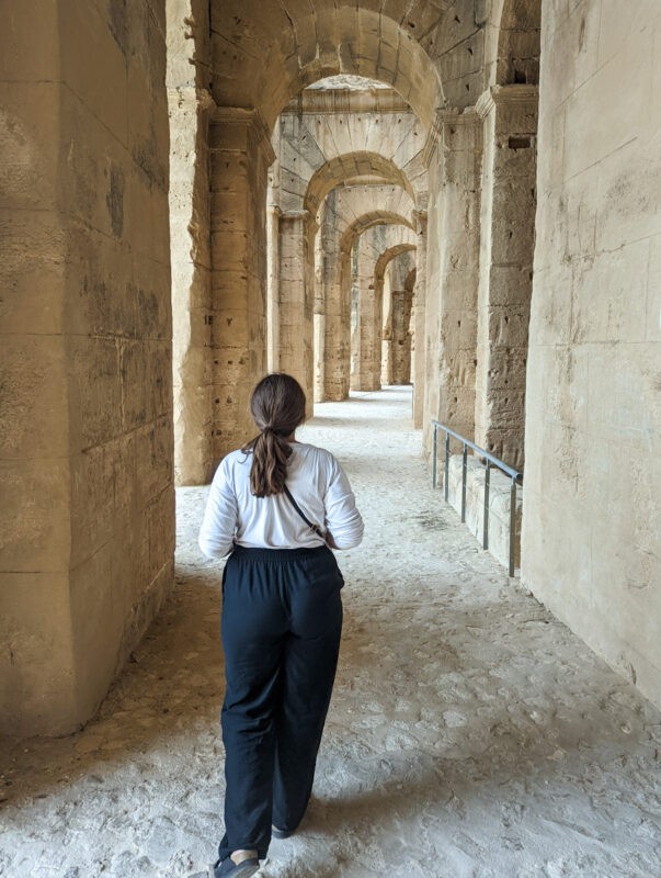 Woman walking through corridors of El Jem wearing a white long sleeved top and black trousers.