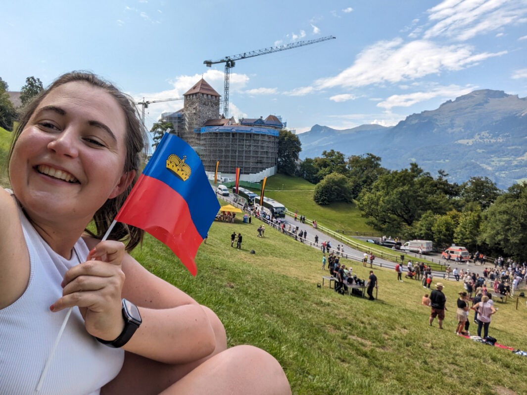 Claire at Vaduz castle with a Liechtenstein flag. She's in the left side of the picture, smiling and holding the flag, and the castle is on the right.