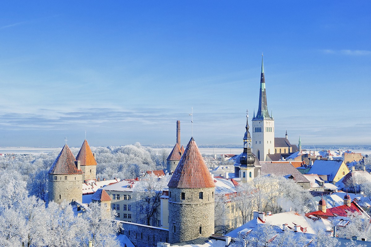 Aerial view of the historic medieval old town of Tallinn covered in snow, showcasing red-roofed buildings, ancient stone towers, and a prominent church spire against a clear blue winter sky