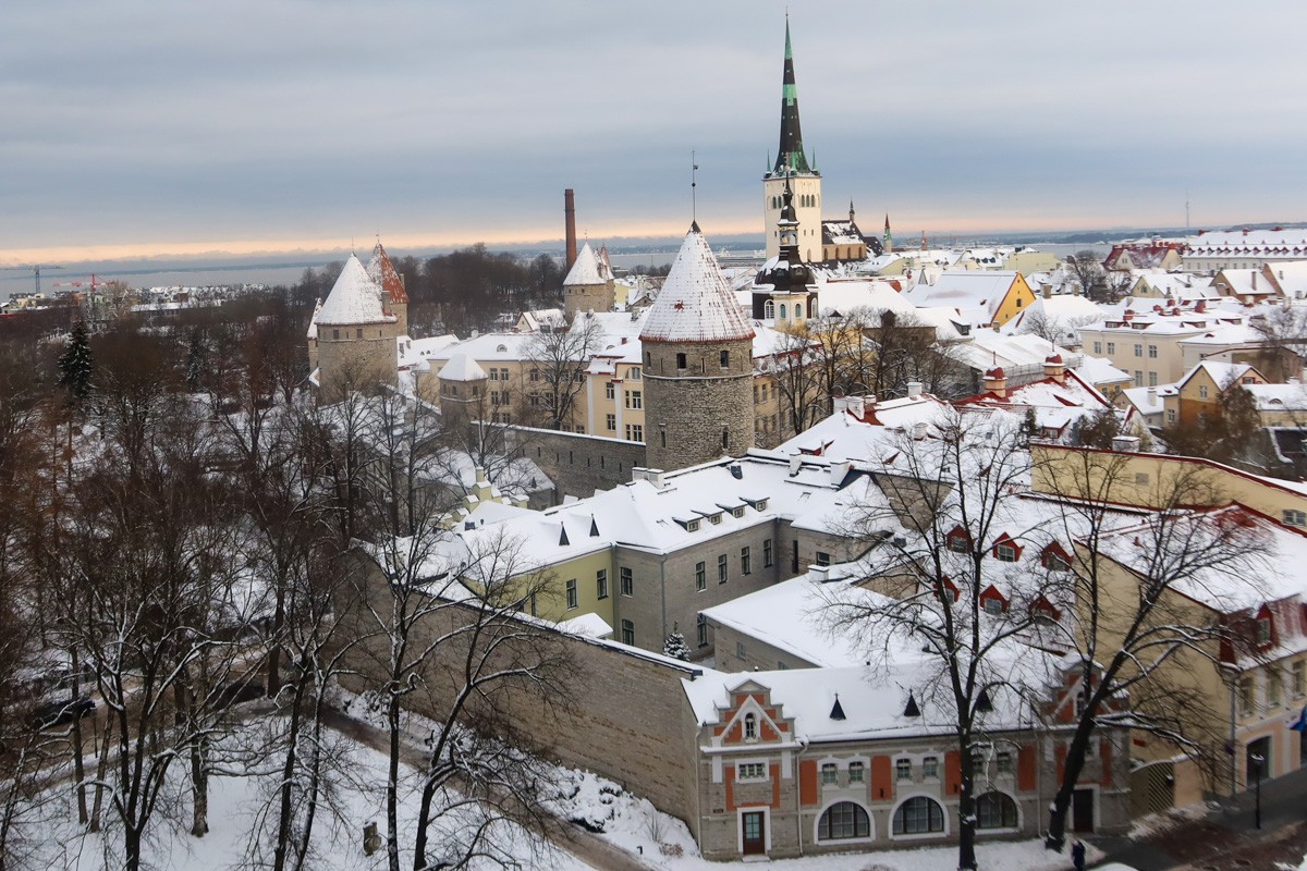 A view of the Old Town of Tallinn, with a church spire, city walls and historic buildings, covered in snow, against a grey sky.