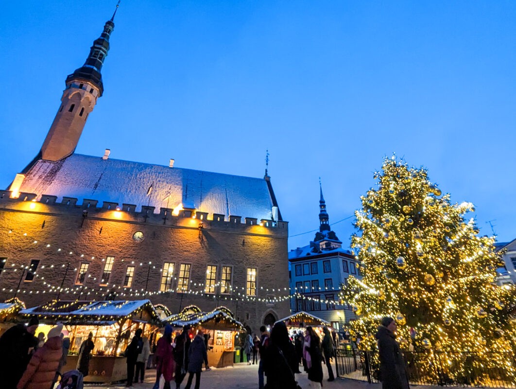 A Christmas market in Tallinn, Estonia, with an illuminated tree and festive lights against the twilight sky, set in front of a grand, historic building.