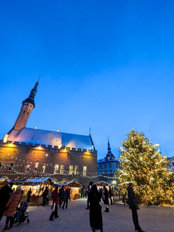 A lively Christmas market scene in Tallinn, Estonia, with an illuminated tree and festive lights against the twilight sky, set in front of the Tallinn Town Hall.