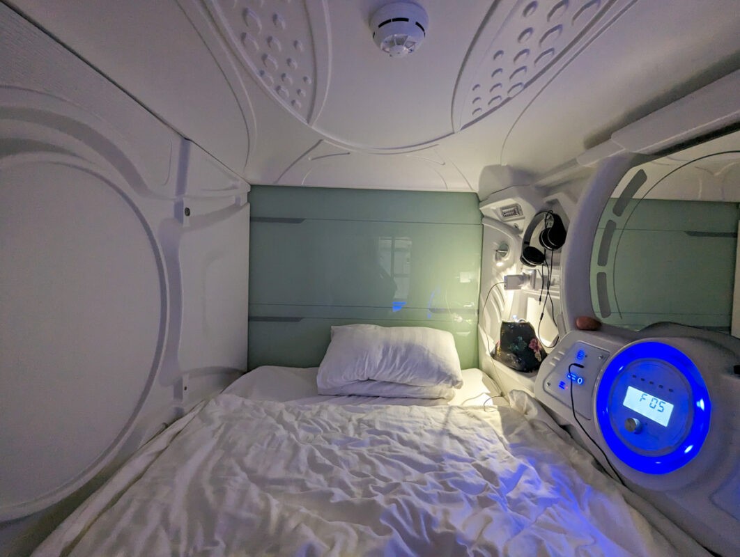 Inside a modern capsule hotel room, featuring a snug bed with white linens and ambient blue lighting, providing a futuristic accommodation experience.