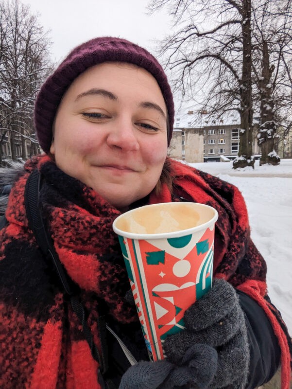 A smiling woman in winter attire holding a festive cup of coffee outdoors, with a snowy Tallinn park in the background, capturing a cheerful moment in the chilly Estonian weather.
