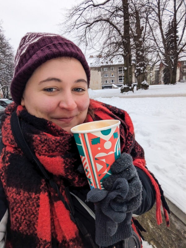 A woman enjoying a warm beverage in a seasonal cup, framed by a wintry Tallinn backdrop, her expression reflecting the cozy charm of the city.