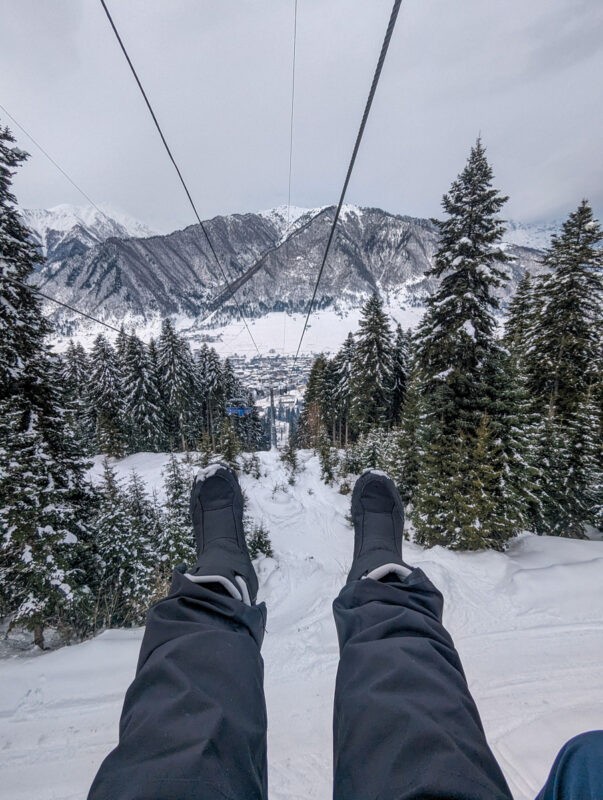 Point of view shot of feet with snow boots on and ski trousers, with snowy trees and mountains.