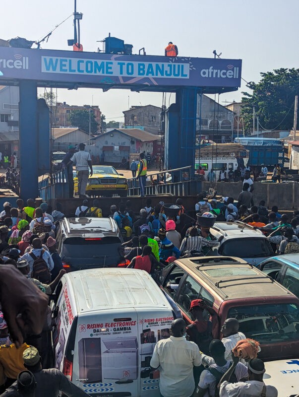 Busy scene at the Banjul ferry terminal with a large crowd of passengers and vehicles waiting to board, under a welcome sign sponsored by Africell.