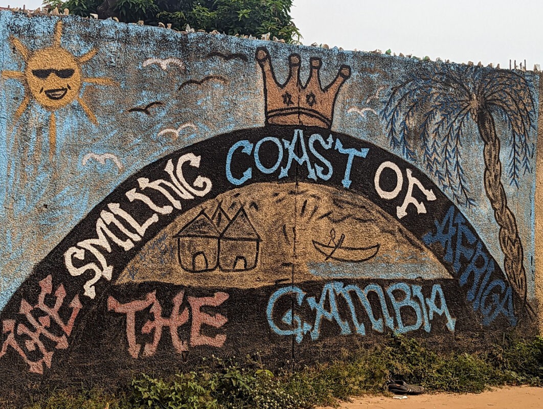 Colorful mural titled 'The Smiling Coast of Africa' with a vibrant depiction of the sun, a crown, palm trees, and houses, celebrating Gambian culture.