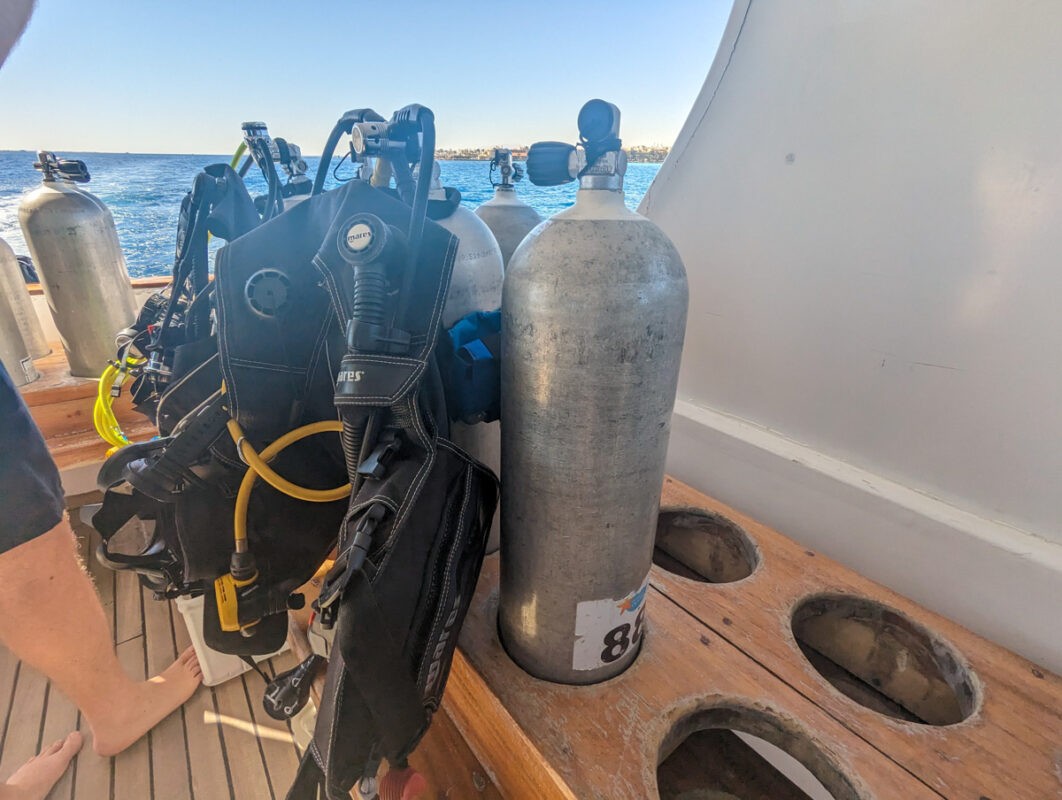 Scuba diving equipment organized on a boat deck, including air tanks, regulators, and BCDs, with a view of the ocean in the background.