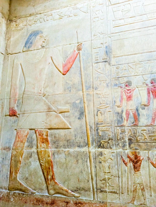 Vivid ancient Egyptian hieroglyphics and carved figures in a range of colors on a temple wall, showing a large standing figure and smaller attendants.
