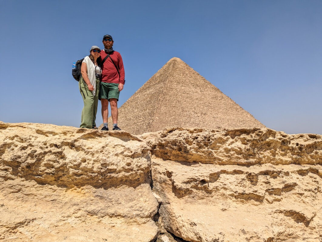 A couple posing for a photo in front of the Great Pyramid of Giza, standing on rocky terrain under a clear blue sky.