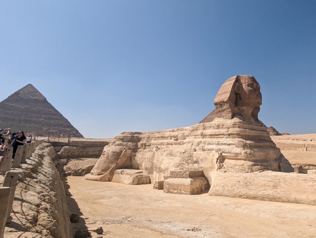 The Great Sphinx of Giza with its limestone body and the Pyramid of Khafre in the background under a clear blue sky, with tourists visible on the left.