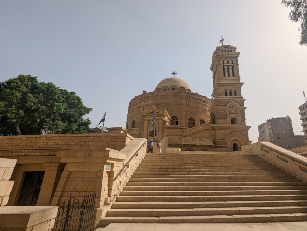 The historic Coptic Orthodox Church of Saint George in Cairo with its rounded dome and prominent bell tower, flanked by stairs and greenery.