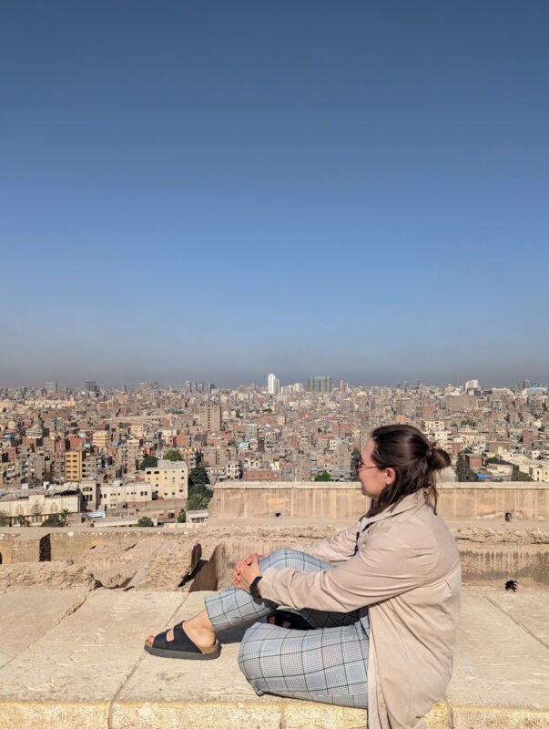 A woman seated on a high ledge overlooking the vast urban landscape of Cairo with numerous buildings stretching to the horizon under a clear sky.