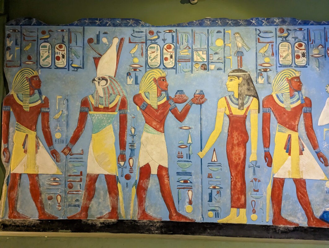 Colorful mural depicting ancient Egyptian figures and hieroglyphics, with four characters holding staffs and offerings in a symmetrical composition.