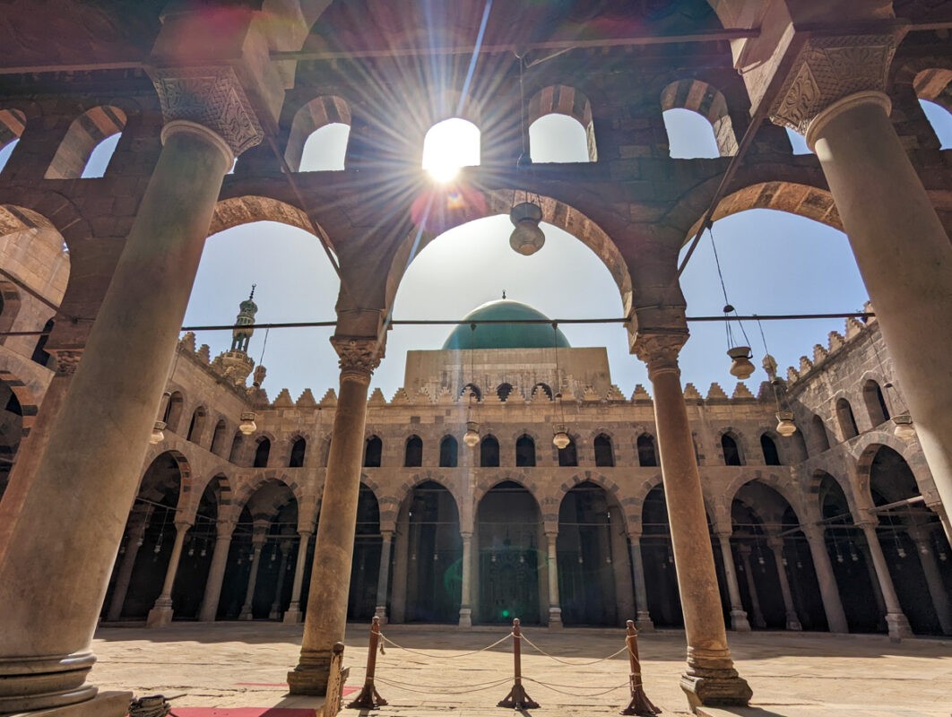 Sunlight streaming through the arches and columns of the Mosque of Ahmad Ibn Tulun in Cairo, highlighting the spacious courtyard and the minaret in the background.