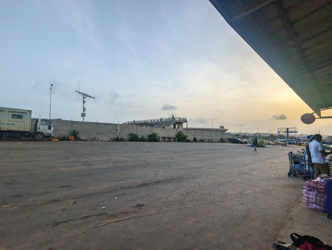 Early morning or late afternoon view at a Senegalese transport hub, with the sun casting a soft glow on the horizon and a few cars and buses in the parking area.