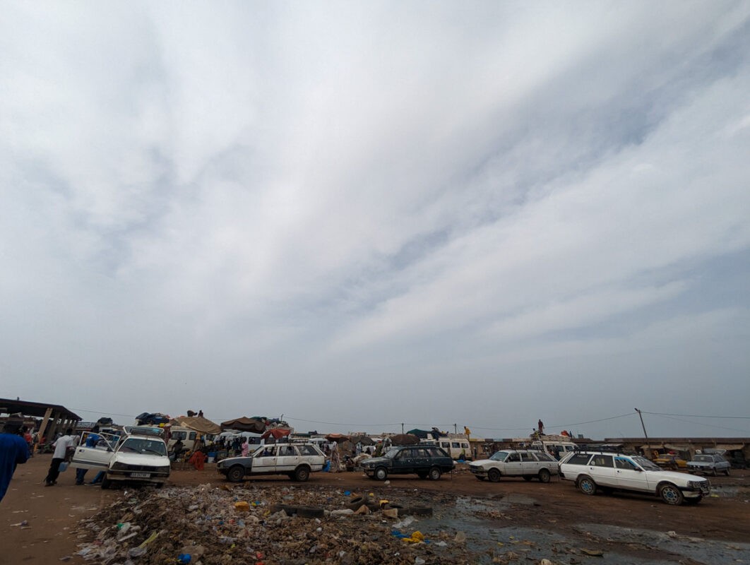 A bustling outdoor scene in Senegal featuring numerous 'sept-places' cars, which are white station wagons loaded with luggage, parked amidst piles of refuse under a hazy sky."