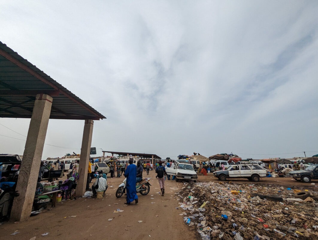 A busy marketplace in Senegal with pedestrians, stalls, and 'sept-places' cars, showcasing a dynamic public transportation hub against a backdrop of overcast skies