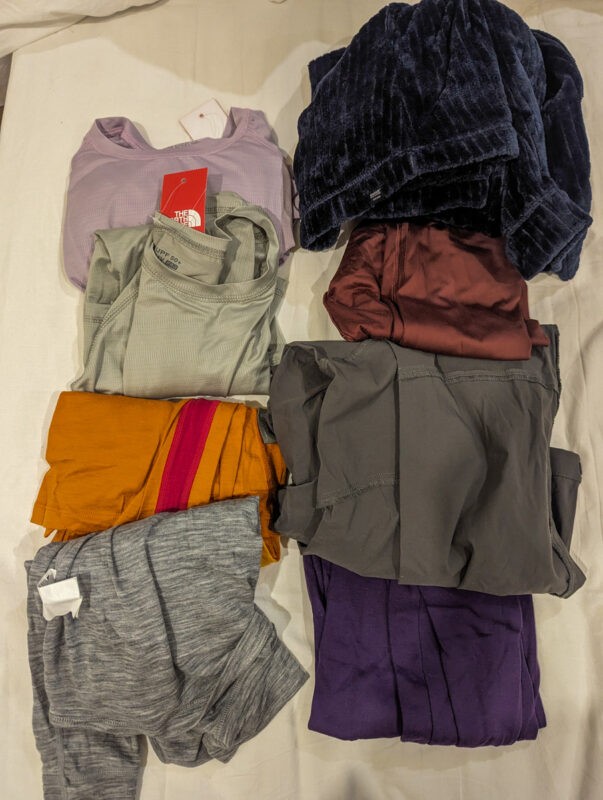 A variety of clothes laid out, including items from The North Face, various tops in shades of purple, grey, orange, and burgundy, along with other clothing items.
