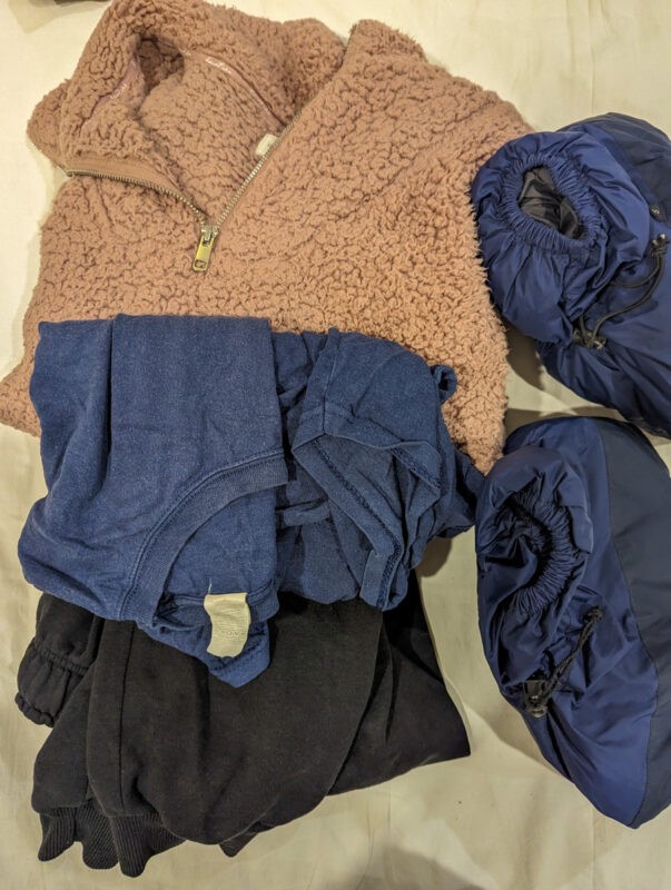 Clothing items arranged, including a pink zip-up fleece, blue and black shirts, and two pairs of blue puffy booties.