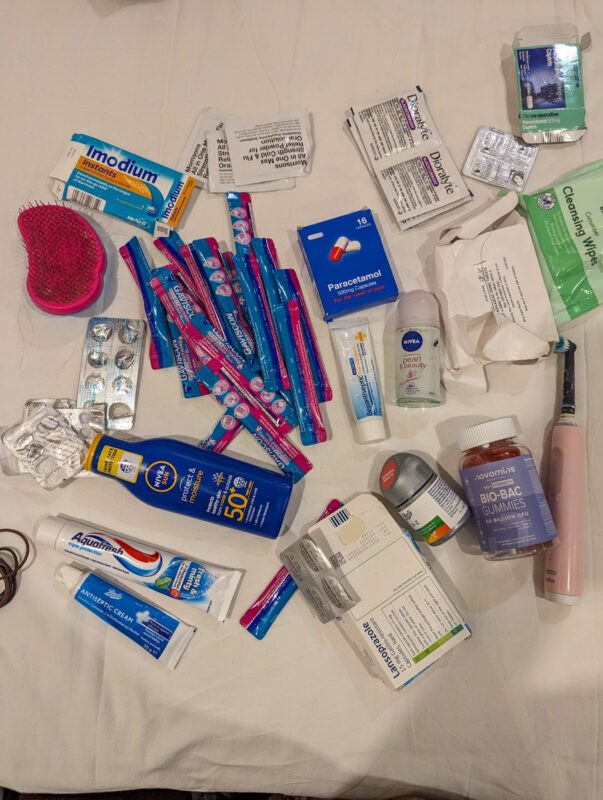 Toiletries and medical supplies scattered on a surface, featuring items like Imodium, paracetamol, cleansing wipes, sunscreen, toothpaste, a toothbrush, and various blister packs.