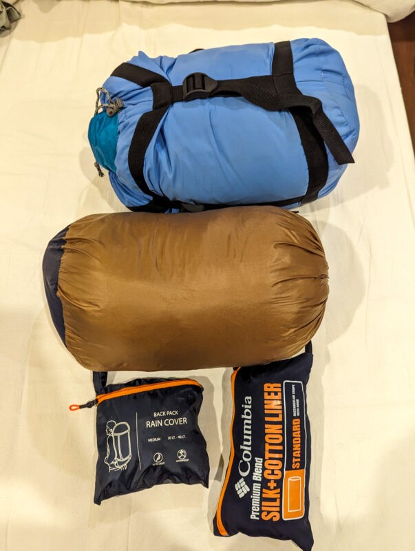 Packed camping gear including a blue sleeping bag, a brown sleeping bag, a rain cover, and a silk-cotton liner, all neatly arranged.