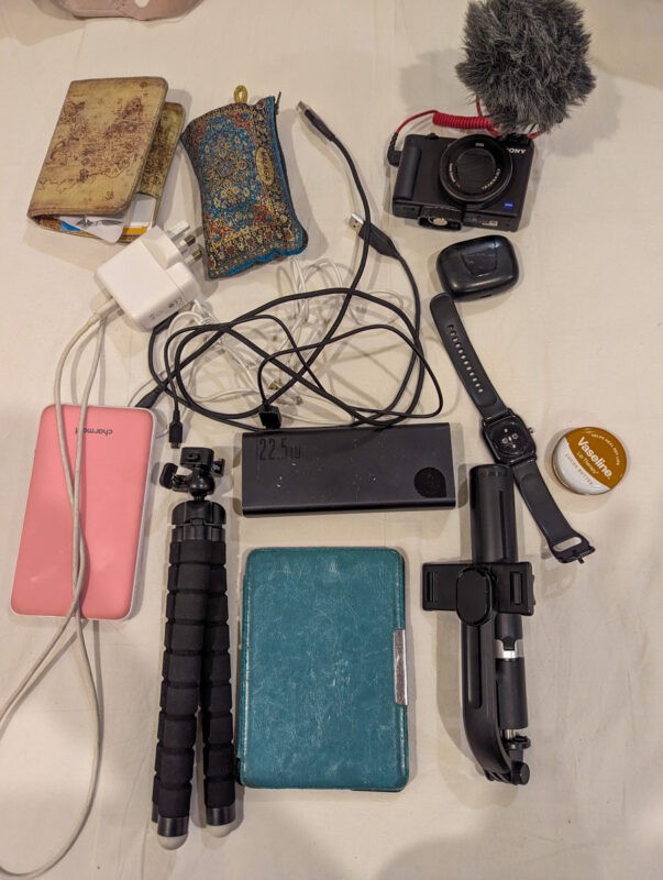 Electronic devices and accessories spread out, including a camera with a microphone, chargers, cables, a tripod, a power bank, a smartwatch, and a teal-colored notebook.