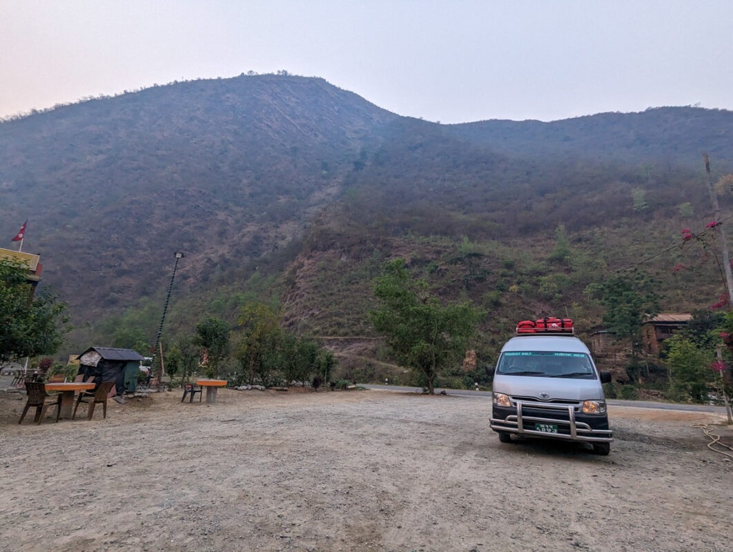 A rugged rural scene in Nepal showing a dusty open space with a parked van, surrounded by hills partially covered with sparse vegetation under a hazy sky.
