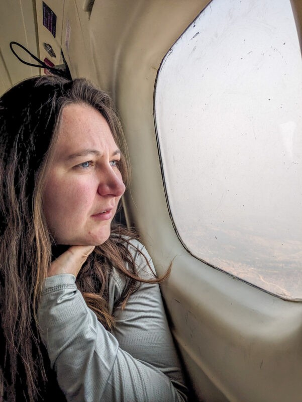 A woman with long brown hair gazes thoughtfully out the window of an airplane, viewing a hazy sky and distant terrain below.