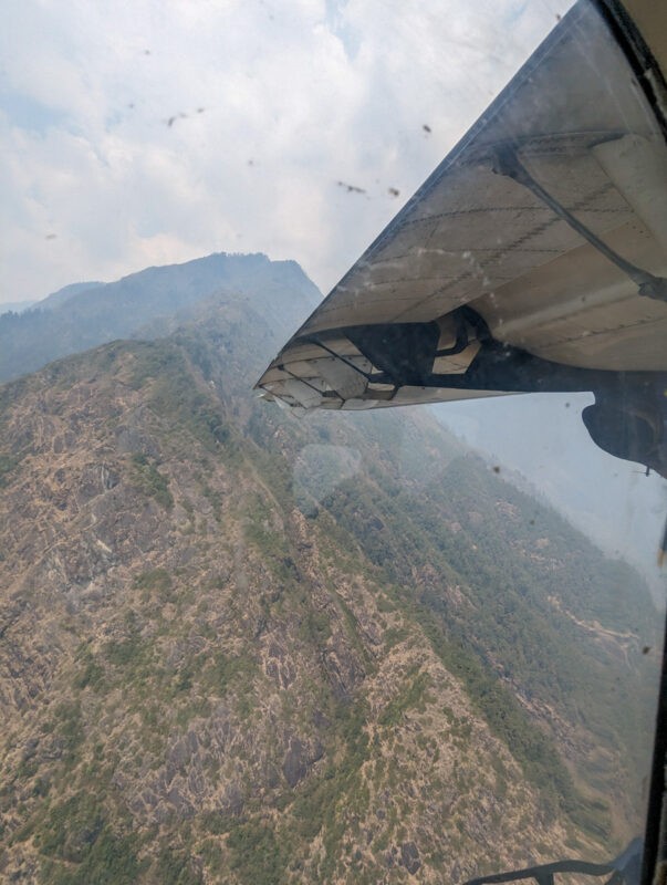 View from an airplane window showing a wing overlooking rugged mountains, with patches of greenery and haze.