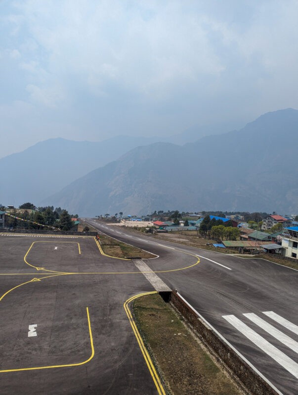 An empty airport runway with yellow lines and markings, surrounded by mountains and a small cluster of buildings under a hazy sky.