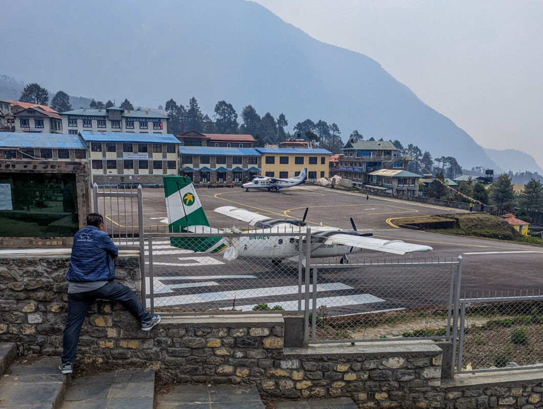 A man wearing a blue jacket leans over a stone fence, watching a small airplane on the tarmac near mountainous terrain.