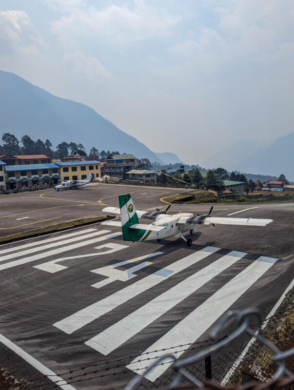 A small airplane with green and white colors on a runway, set against a backdrop of mountains and a clear sky.