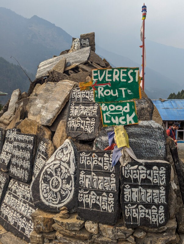 A pile of Mani stones painted with Buddhist mantras, including a sign that reads "Everest Route Good Luck," set against a mountainous backdrop.