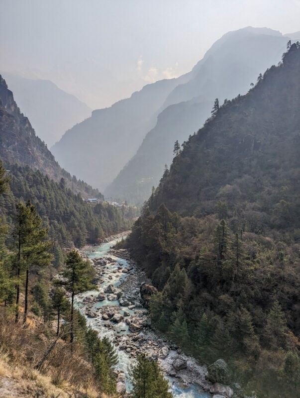 A river flows through a mountainous landscape with pine trees, under hazy skies with distant peaks barely visible.
