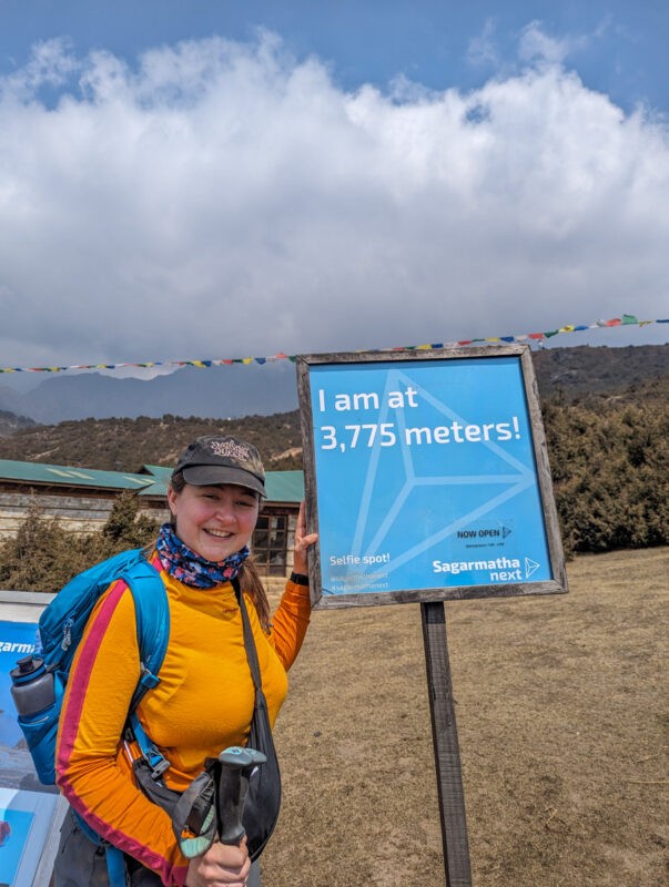  female hiker smiling beside an altitude marker sign at 3,775 meters, surrounded by a mountain landscape under a partly cloudy sky.
