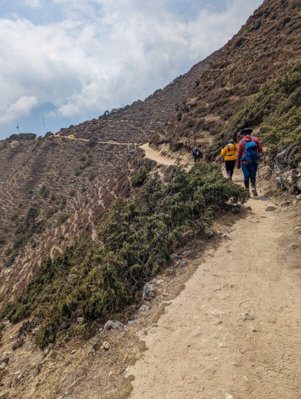 A trail winds along a mountainside with sparse vegetation and hikers walking in the distance, under a partly cloudy sky