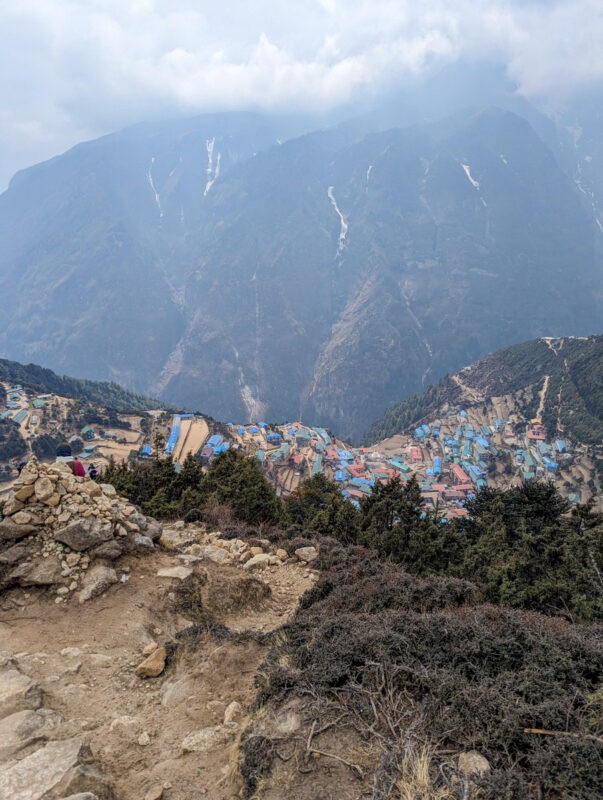 Elevated view of a colorful mountain village nestled in a deep valley, with rugged mountains shrouded in clouds in the distance.