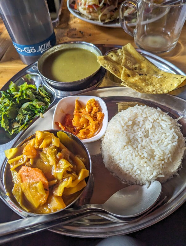 A plate of dal baht, with rice, curry, soup, greens, poppadom and pickles.