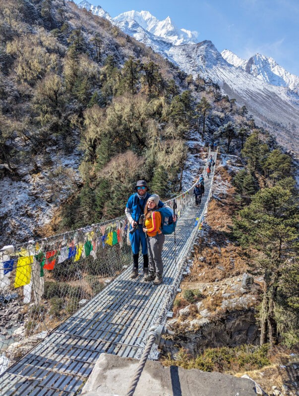 Two hikers, one male and one female, cross a suspension bridge decorated with prayer flags, surrounded by forested hills and a snowy mountain backdrop