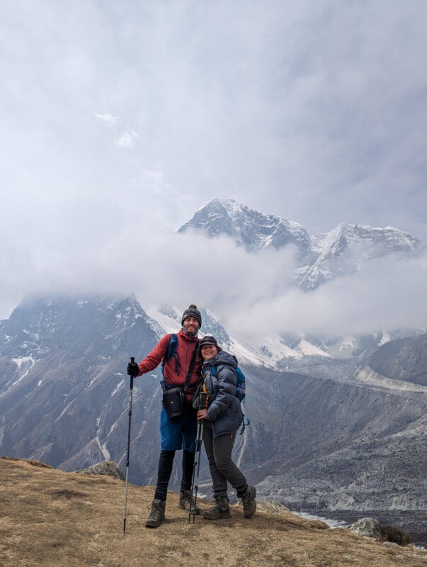 Two hikers, one male and one female, smile at the camera with trekking poles in hand, backed by partially cloud-covered mountains.
