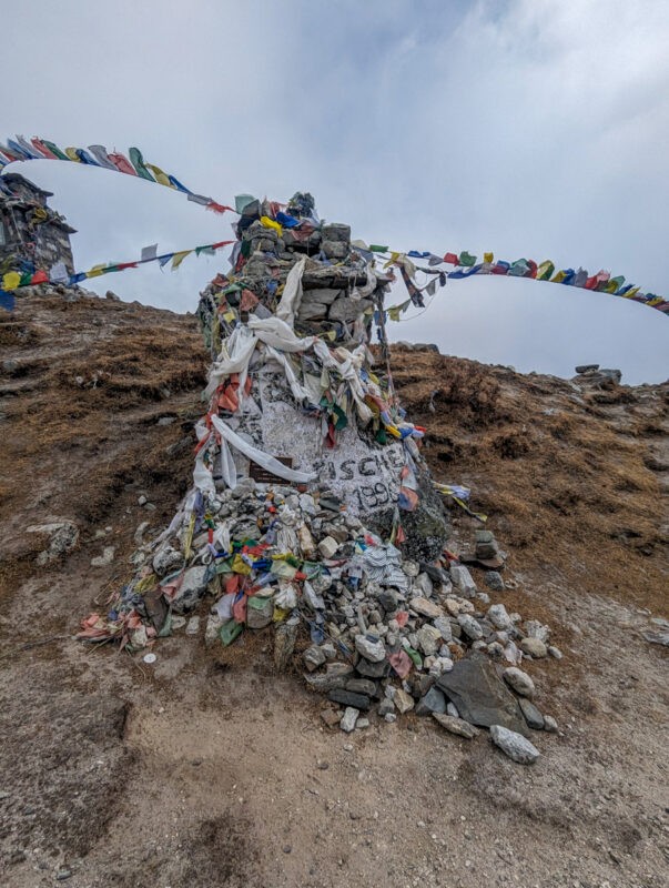 A rock cairn topped with colorful prayer flags on a mountain ridge, surrounded by rocky terrain and a faint view of distant mountains.