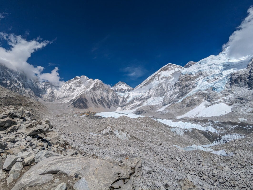 A panoramic view of the Everest Base Camp area showing rocky terrain, snow, and part of a glacial area under a clear blue sky.