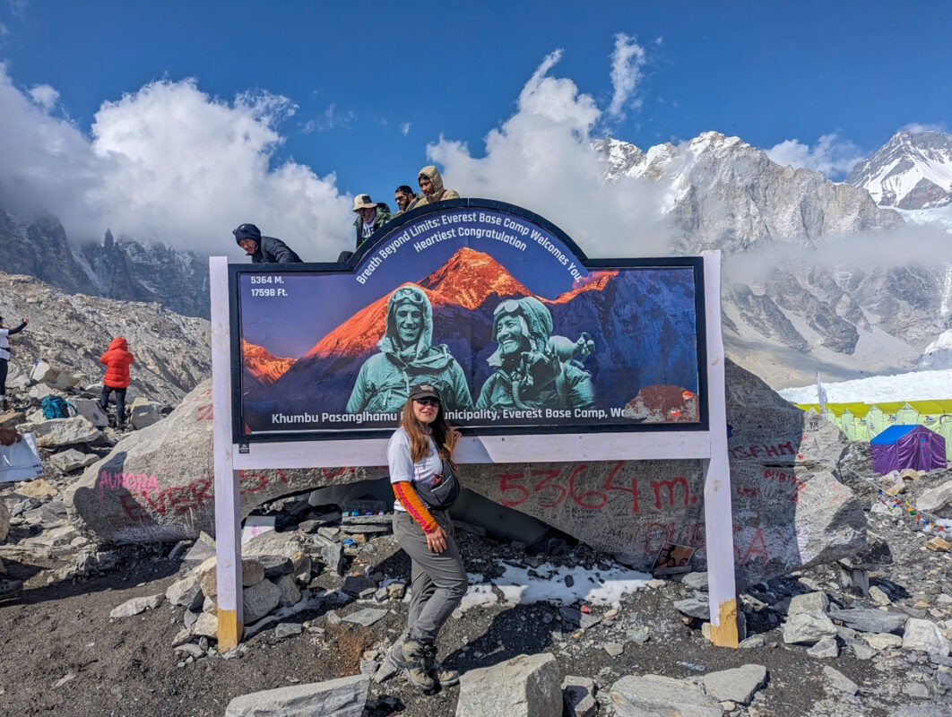 A person stands in front of the Everest Base Camp welcome sign at 5364 meters elevation, surrounded by rocky terrain and snowy mountain peaks under a clear blue sky