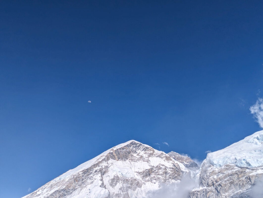 The moon visible in a clear blue sky above the snow-covered summit of a towering mountain.