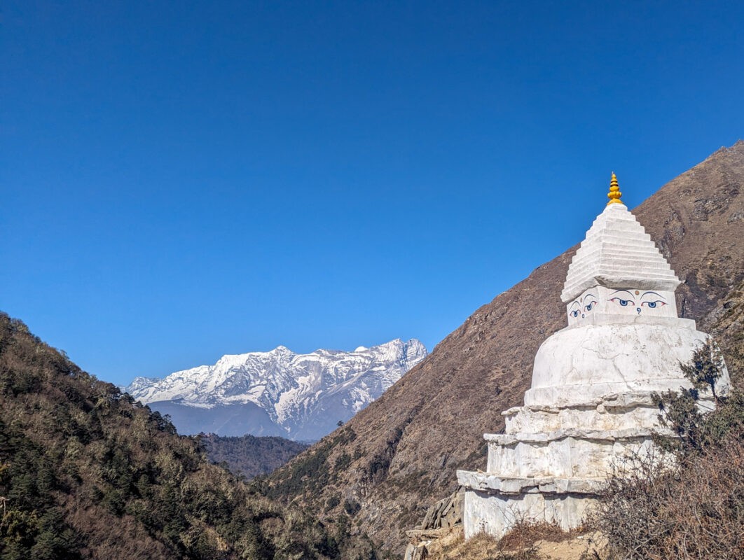 A large, white Buddhist stupa decorated with painted eyes, standing against a mountainous backdrop with snowy peaks under a clear blue sky.