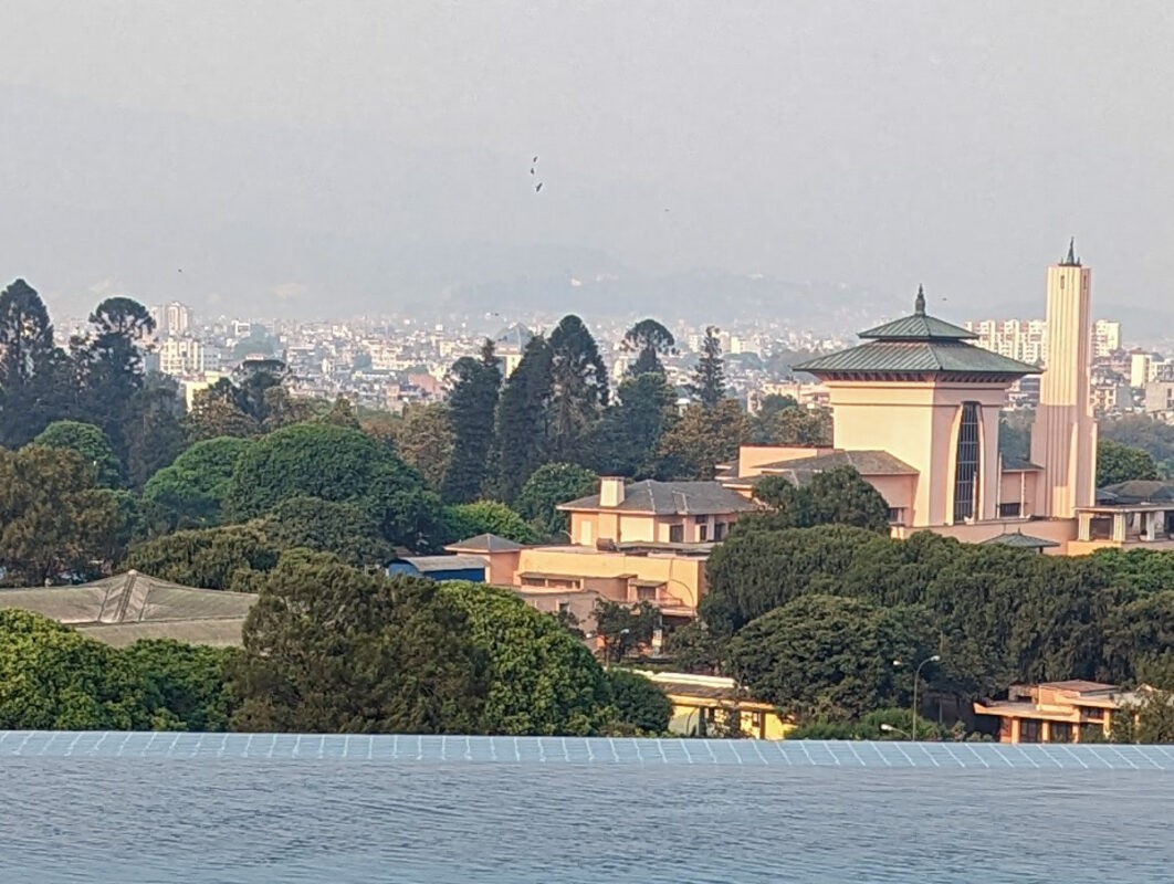 Panoramic view overlooking a lush green park and a large water body in the foreground, with an iconic building featuring a unique dome and tall, slender tower, set against a cityscape backdrop under a clear sky.